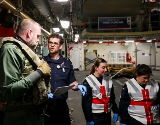 Team of workers in a shipyard or aircraft carrier hangar discussing work, with one officer in navy uniform and two women in reflective vests and gloves.