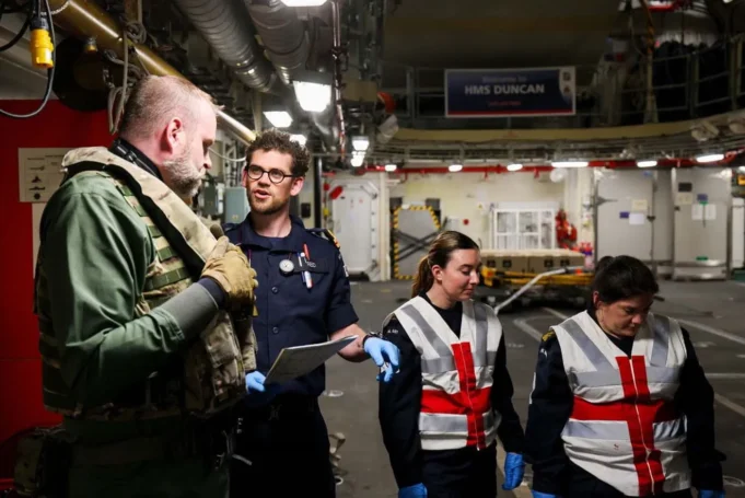 Inside the dramatic carrier air ambulance rescue at sea Team of workers in a shipyard or aircraft carrier hangar discussing work, with one officer in navy uniform and two women in reflective vests and gloves.