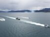 Royal Navy tests killer drone-helicopter team in Norway Several boats sail in formation across a calm sea, leaving white wake trails with a mountainous coastline and overcast sky in the background.