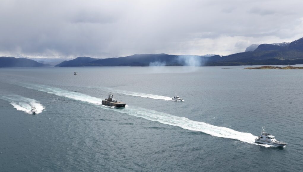 FLEET-20260511-ZC0198-013 Several boats sail in formation across a calm sea, leaving white wake trails with a mountainous coastline and overcast sky in the background.