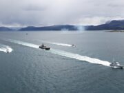 Several boats sail in formation across a calm sea, leaving white wake trails with a mountainous coastline and overcast sky in the background.