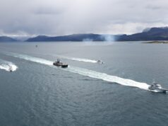 Several boats sail in formation across a calm sea, leaving white wake trails with a mountainous coastline and overcast sky in the background.