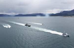 Royal Navy tests killer drone-helicopter team in Norway Several boats sail in formation across a calm sea, leaving white wake trails with a mountainous coastline and overcast sky in the background.