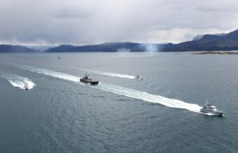 Several boats sail in formation across a calm sea, leaving white wake trails with a mountainous coastline and overcast sky in the background.