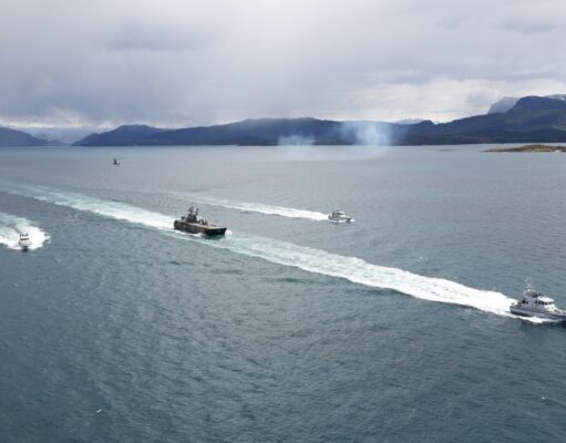 Several boats sail in formation across a calm sea, leaving white wake trails with a mountainous coastline and overcast sky in the background.