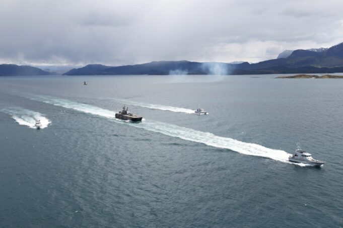 Royal Navy tests killer drone-helicopter team in Norway Several boats sail in formation across a calm sea, leaving white wake trails with a mountainous coastline and overcast sky in the background.