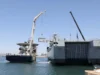 HMS Stirling Castle delivers kit for Hormuz mission Two large ships at a harbor with cranes lifting a container between them, likely a transfer operation at sea.