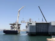 Two large ships at a harbor with cranes lifting a container between them, likely a transfer operation at sea.