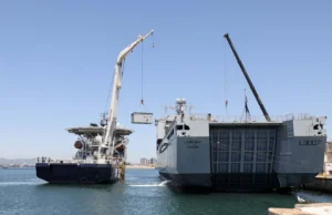 Two large ships at a harbor with cranes lifting a container between them, likely a transfer operation at sea.