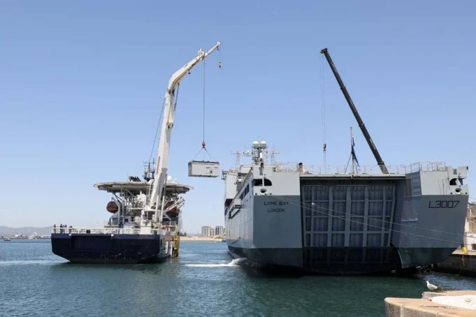 HMS Stirling Castle delivers kit for Hormuz mission Two large ships at a harbor with cranes lifting a container between them, likely a transfer operation at sea.
