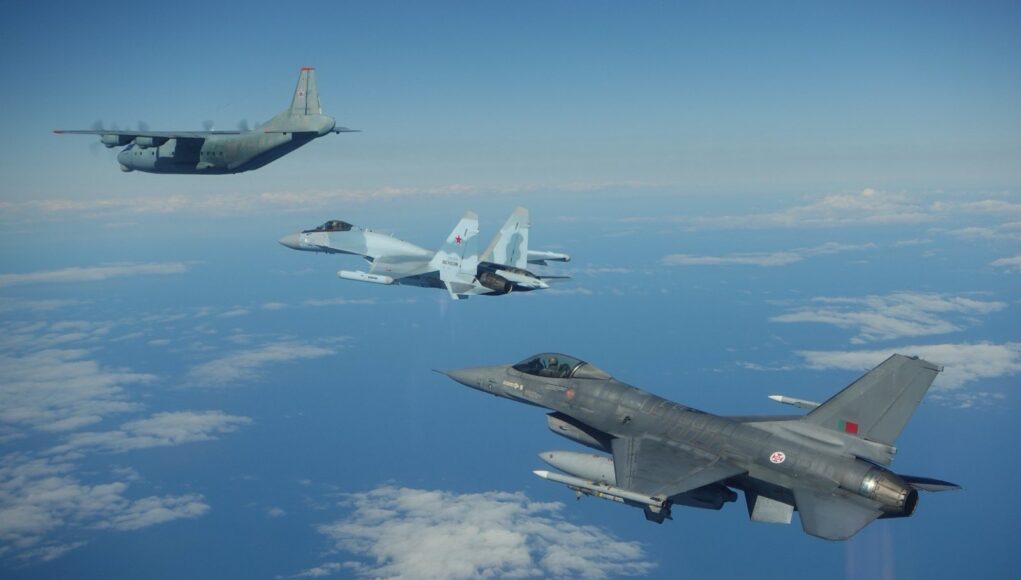 Three military aircraft in formation over the ocean with scattered clouds beneath them.