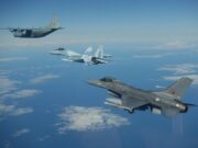 Three military aircraft in formation over the ocean with scattered clouds beneath them.