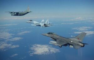 Three military aircraft in formation over the ocean with scattered clouds beneath them.