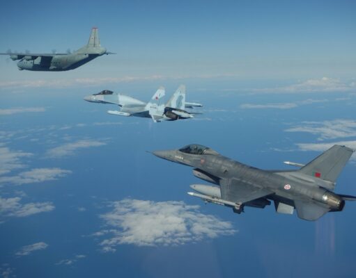 Three military aircraft in formation over the ocean with scattered clouds beneath them.