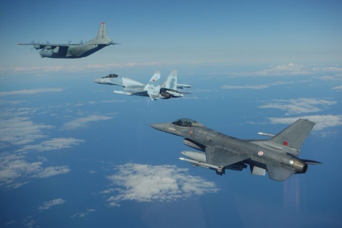 Three military aircraft in formation over the ocean with scattered clouds beneath them.