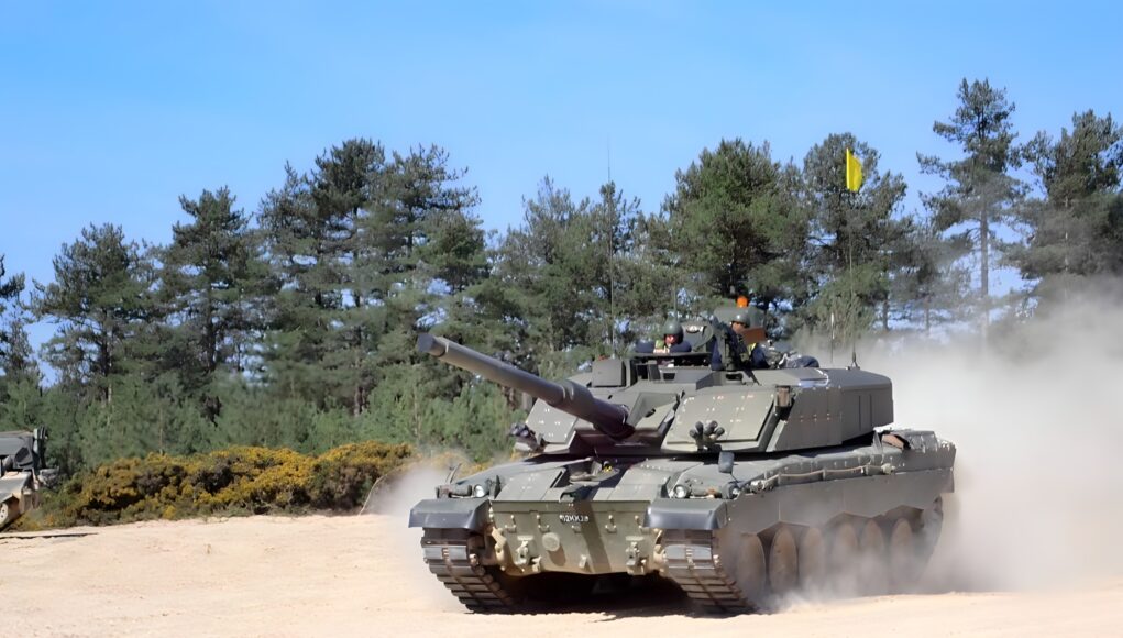 Armored tank moves across a dusty field with trees behind it and a blue sky above; smoke rises from the tracks as it advances.