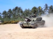 Britain’s new battle tank completes mission trials Armored tank moves across a dusty field with trees behind it and a blue sky above; smoke rises from the tracks as it advances.
