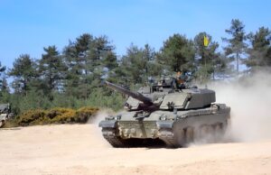 Armored tank moves across a dusty field with trees behind it and a blue sky above; smoke rises from the tracks as it advances.