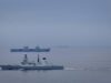 Large gray naval warship in the foreground on calm water, with smaller ships visible on the horizon (hull marked D37).