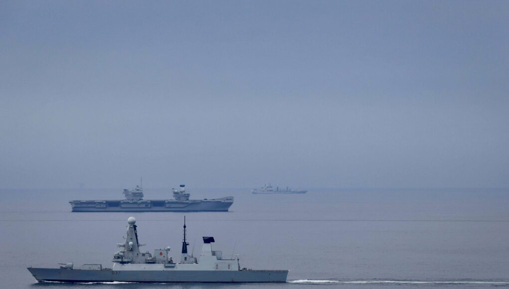 Large gray naval warship in the foreground on calm water, with smaller ships visible on the horizon (hull marked D37).