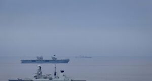 Large gray naval warship in the foreground on calm water, with smaller ships visible on the horizon (hull marked D37).
