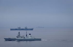 Large gray naval warship in the foreground on calm water, with smaller ships visible on the horizon (hull marked D37).