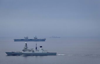 Large gray naval warship in the foreground on calm water, with smaller ships visible on the horizon (hull marked D37).