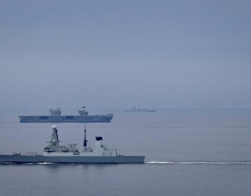 Destroyer joins aircraft carrier for Nordic deployment Large gray naval warship in the foreground on calm water, with smaller ships visible on the horizon (hull marked D37).