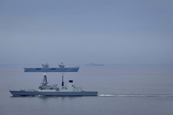 Large gray naval warship in the foreground on calm water, with smaller ships visible on the horizon (hull marked D37).