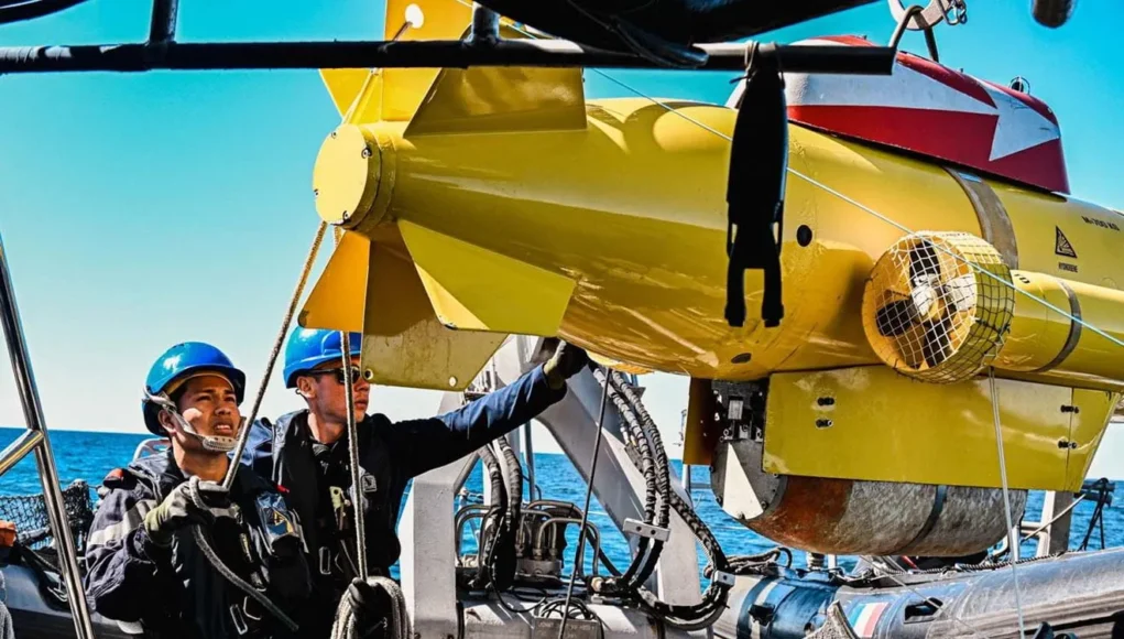 HISXLrUXgAA0X7x Two crew members in blue hard hats on a boat lowering a yellow submarine-like ROV into the sea.