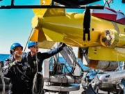 Two crew members in blue hard hats on a boat lowering a yellow submarine-like ROV into the sea.