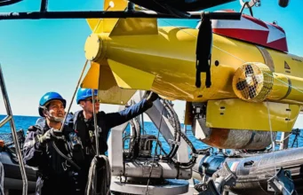 Two crew members in blue hard hats on a boat lowering a yellow submarine-like ROV into the sea.
