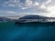 Submarine partially submerged with the hull just below the surface and a periscope rising above the water under a cloudy sky.