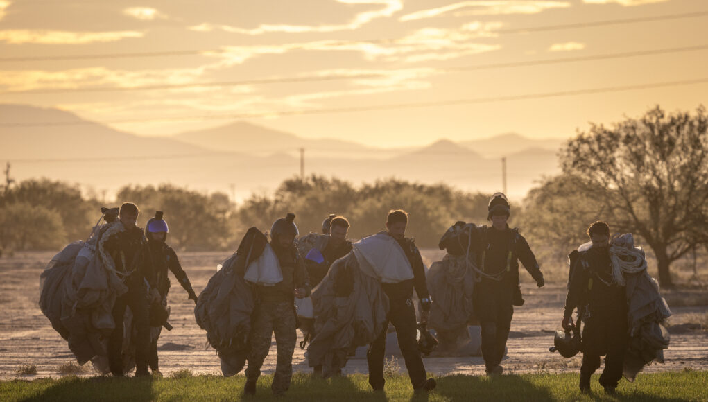 Group of skydivers in full gear and parachutes walking together across a field at sunset with mountains in the distance at the horizon.