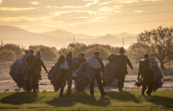 Group of skydivers in full gear and parachutes walking together across a field at sunset with mountains in the distance at the horizon.