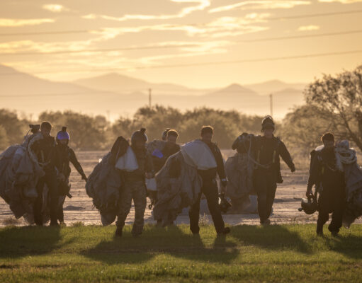 Group of skydivers in full gear and parachutes walking together across a field at sunset with mountains in the distance at the horizon.