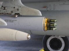 Close-up of a wing-mounted rocket launcher under a military aircraft, showing yellow-tipped missiles ready for launch