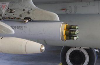 Close-up of a wing-mounted rocket launcher under a military aircraft, showing yellow-tipped missiles ready for launch
