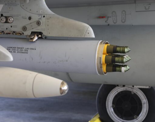 Close-up of a wing-mounted rocket launcher under a military aircraft, showing yellow-tipped missiles ready for launch