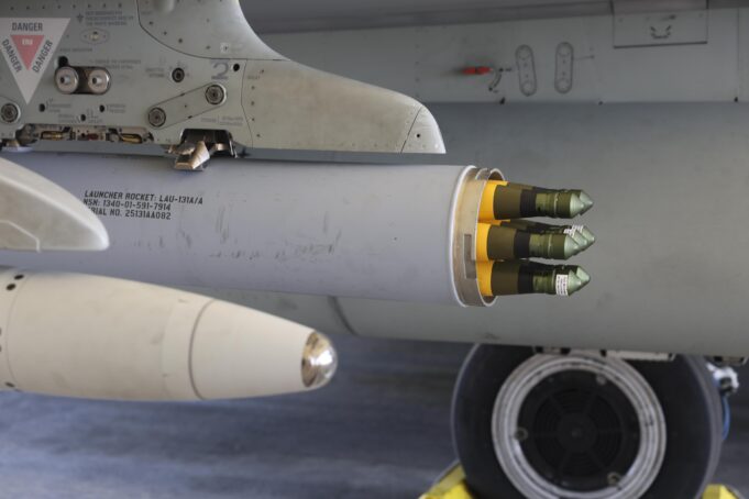 Close-up of a wing-mounted rocket launcher under a military aircraft, showing yellow-tipped missiles ready for launch