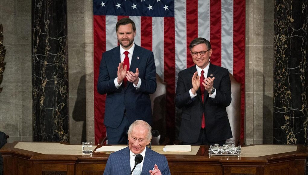 A man at a wooden podium speaking into a microphone with two suited men applauding behind him, in front of a large U.S. flag backdrop.
