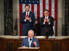 A man at a wooden podium speaking into a microphone with two suited men applauding behind him, in front of a large U.S. flag backdrop.