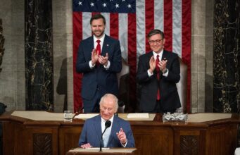 King Charles plays a blinder but will it move the dial? A man at a wooden podium speaking into a microphone with two suited men applauding behind him, in front of a large U.S. flag backdrop.