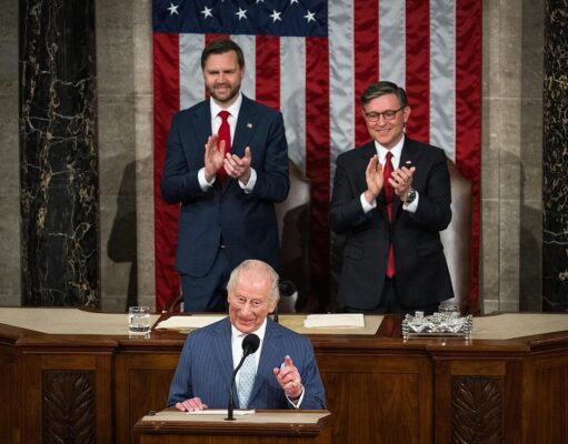 A man at a wooden podium speaking into a microphone with two suited men applauding behind him, in front of a large U.S. flag backdrop.