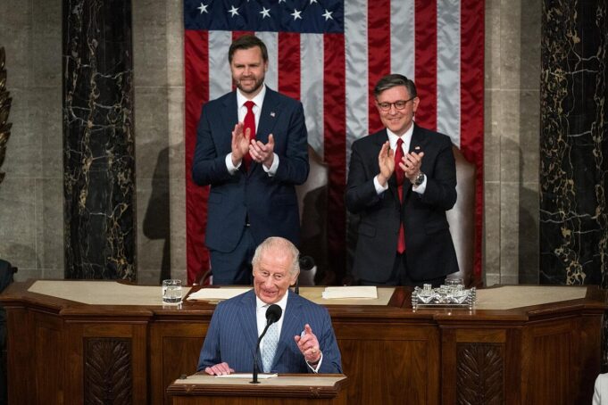 A man at a wooden podium speaking into a microphone with two suited men applauding behind him, in front of a large U.S. flag backdrop.