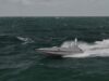 Gray speedboat racing across choppy dark green ocean with white spray at the stern, under a cloudy sky.