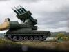 Large green mobile missile launcher with four missiles raised on a field under a cloudy sky.