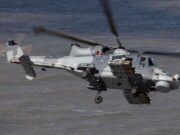 Grey military helicopter in flight over water, with rockets mounted on its side and rotor blur indicating motion.