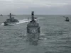 Three gray military ships sail in formation on the open sea, with a helicopter hovering to the right.