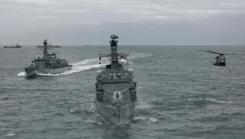 Three gray military ships sail in formation on the open sea, with a helicopter hovering to the right.